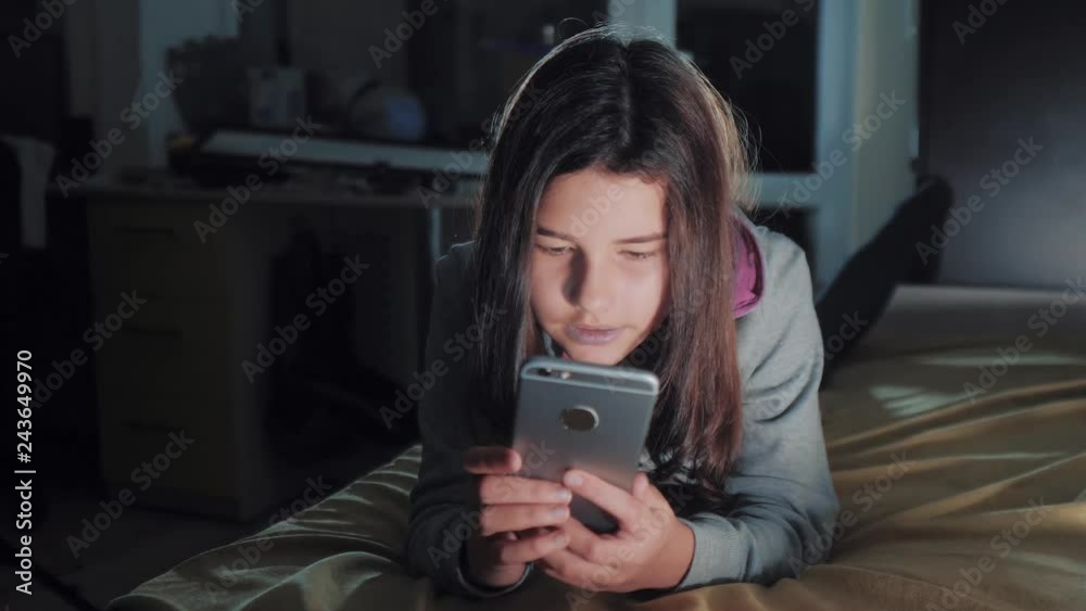 Teenage girl online shopping looks on her smartphone in bed during the night. little teen hood girl writes a message chatting in social media lifestyle messenger night evening indoors smartphone on Teenage girl online shopping looks on her smartphone in bed during the night. little teen hood girl writes a message chatting in social media lifestyle messenger night evening indoors smartphone on