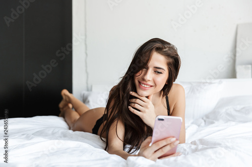 Fotografie Beautiful brunette woman wearing lingerie using mobile phone talking lies in bed