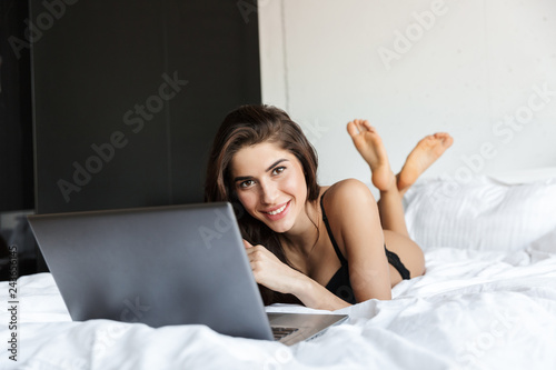 Beautiful brunette woman wearing lingerie using laptop computer lies in bed.