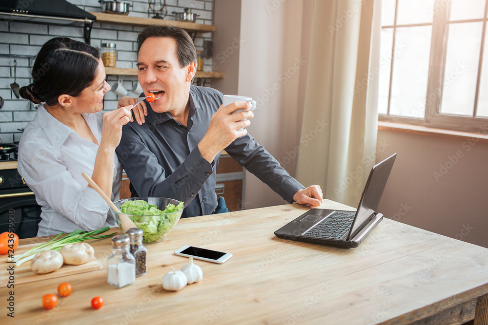 Fototapeta premium Cheerful people sit together in room. Woman feed guy with tometo. He keep moouth opened. Man hold white cup.