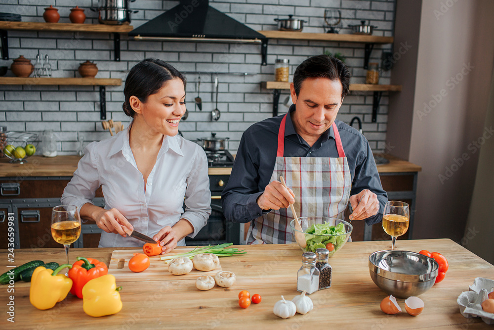 Man and woman sit together at table in kitchen. They prepare dinner together. Woman cut tomato and look at husband. He blend salad in glass bown.