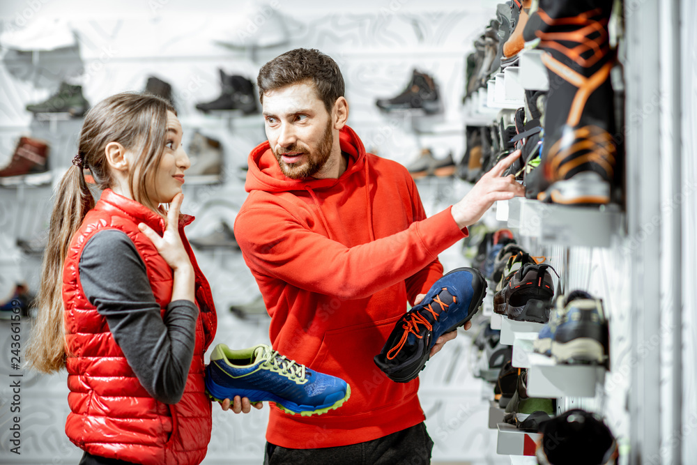 Fototapeta premium Mam and woman in red sports clothes choosing trail shoes for hiking standing near the showacase of the modern sports shop