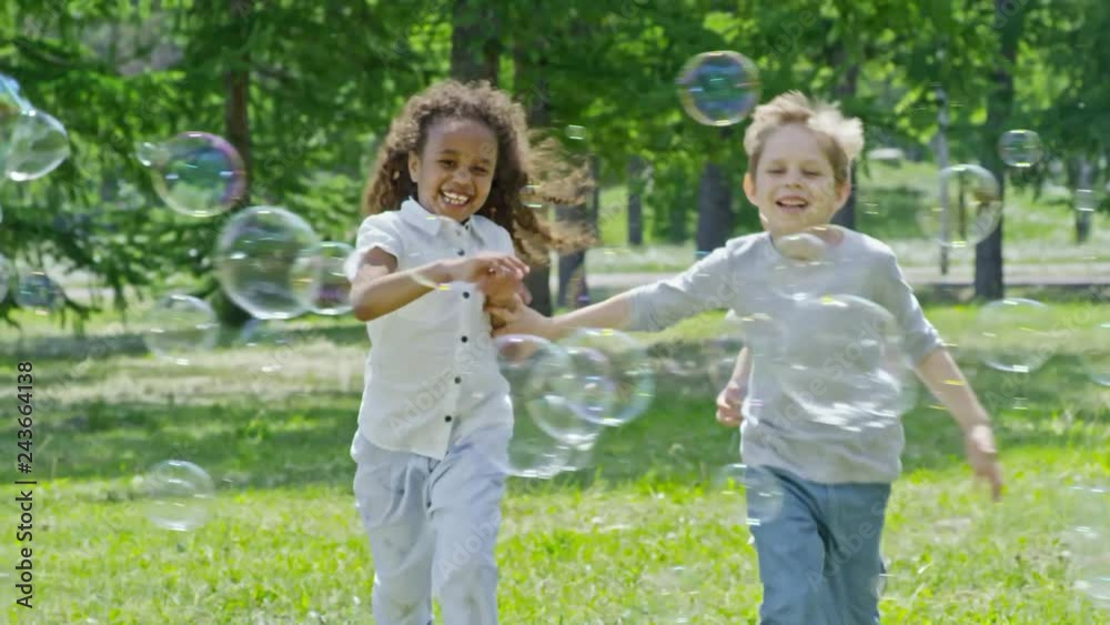 Little friends laughing and running on green lawn in the park through lots of flying soap bubbles. Footage in slowmo Little friends laughing and running on green lawn in the park through lots of flying soap bubbles. Footage in slowmo