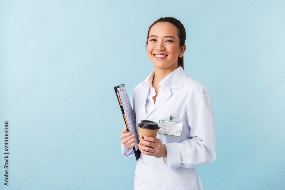 Woman doctor posing isolated over blue wall background holding clipboard and drinking coffee. Woman doctor posing isolated over blue wall background holding clipboard and drinking coffee.