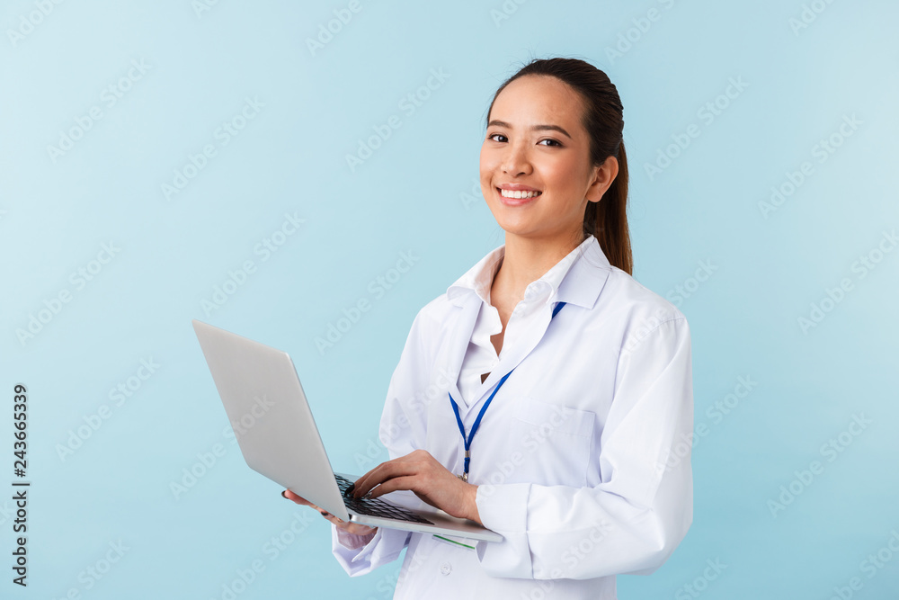 Young woman doctor posing isolated over blue wall background using laptop computer. Young woman doctor posing isolated over blue wall background using laptop computer.