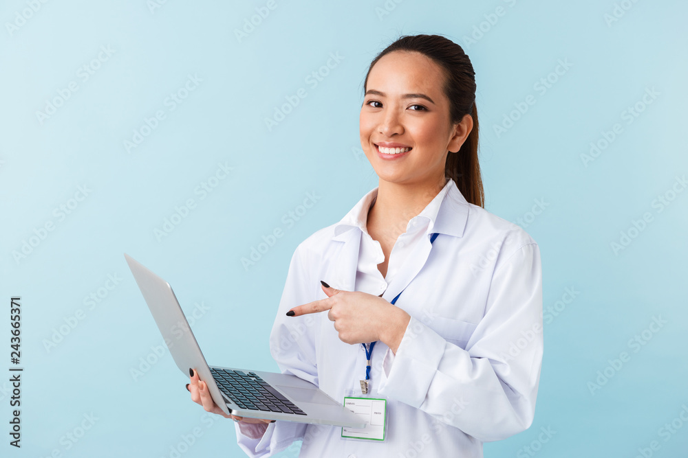 Young woman doctor posing isolated over blue wall background using laptop computer. Young woman doctor posing isolated over blue wall background using laptop computer.