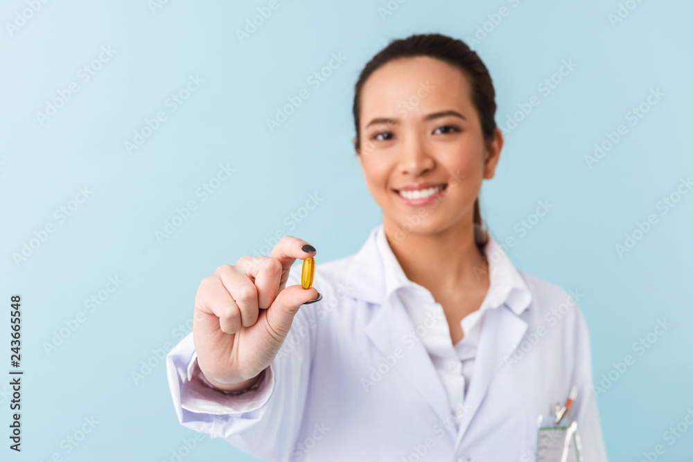 Young woman doctor posing isolated over blue wall background holding pills. Young woman doctor posing isolated over blue wall background holding pills.