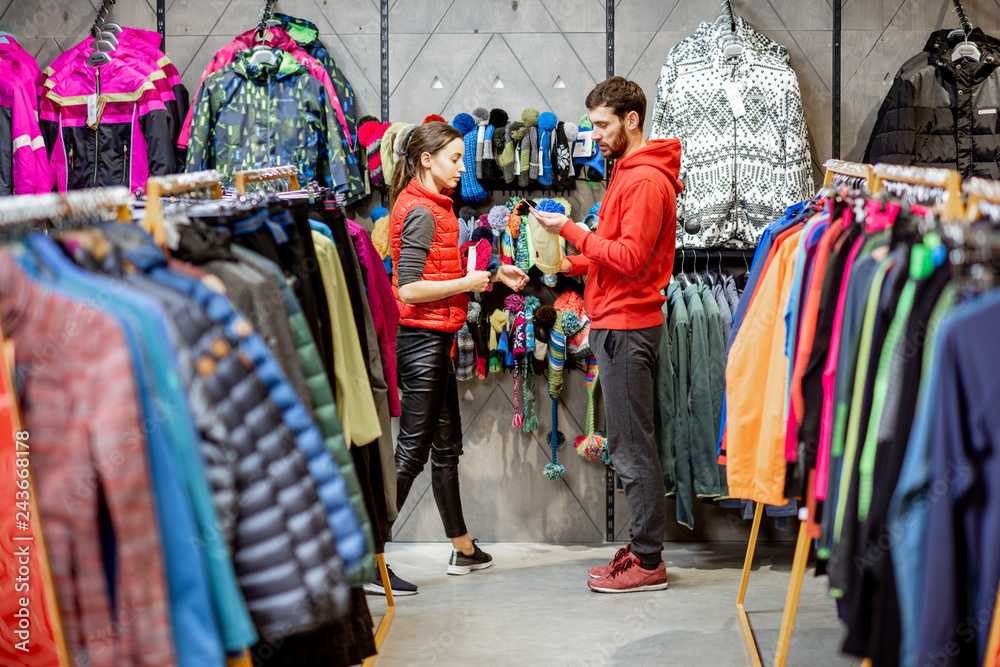 Obraz premium Man and woman looking for winter clothes standing together in the sports shop with colorful jackets and garment