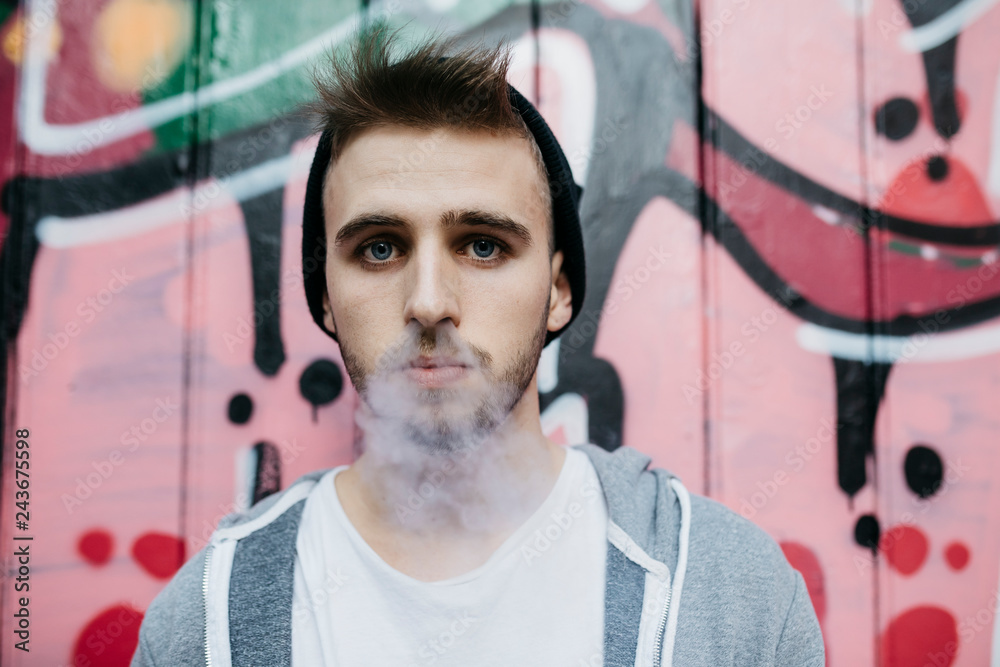 Young man standing in front of graffiti, smoking electronic cigarette