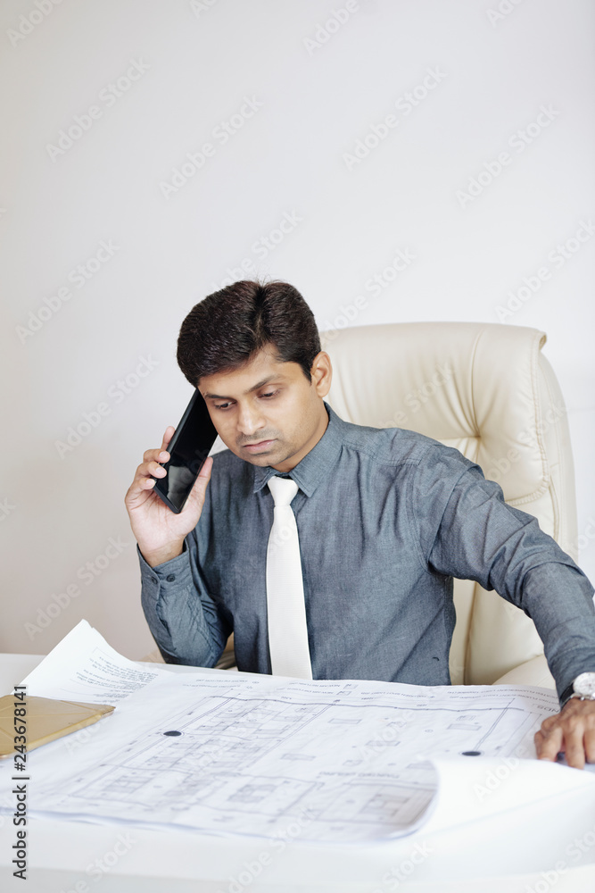 Serious Indian architect checking constuction plan of building and talking on phone Serious Indian architect checking constuction plan of building and talking on phone