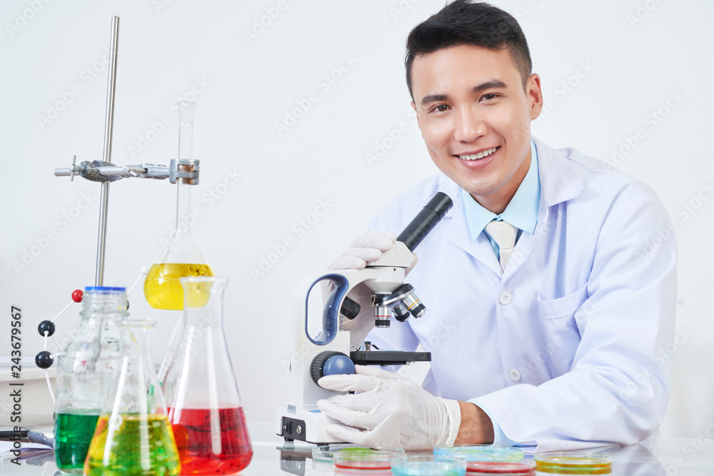 Portrait of happy young Vietnamese chemist looking at various petri dishes through microscope Portrait of happy young Vietnamese chemist looking at various petri dishes through microscope
