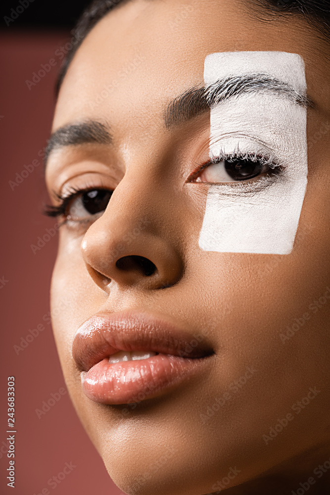 close-up view of attractive african american girl with white paint stroke on eye looking at camera isolated on brown
