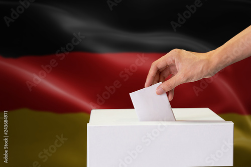 Close-up of human hand casting and inserting a vote and choosing and making a decision what he wants in polling box with Germany flag blended in background.