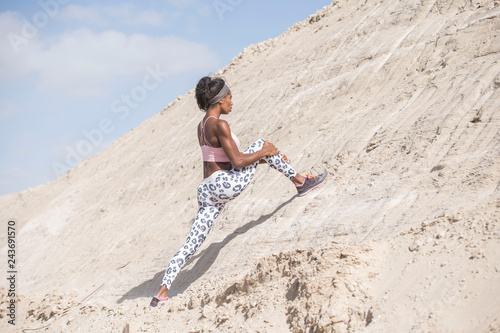 Strong athletic African American Black woman wearing long black and white printed tights and a pink sports bra is stretching on a dusty rocky desert hillside 