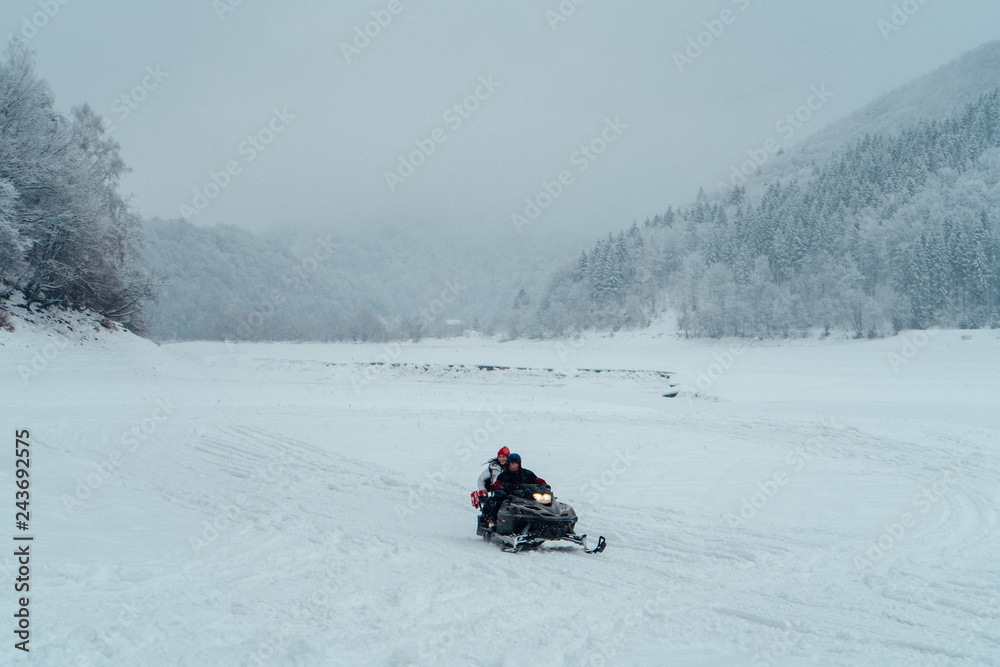 Man and woman riding on a snowmobile on the frozen lake in the mountains with the scenic view. Pine trees covered with snow