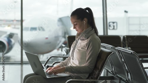 Girl freelancer with headphones in ears using laptop for remote work and waiting her flight at international airport
