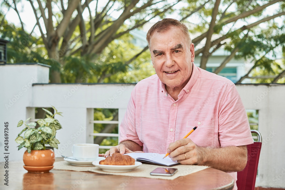 Aged smiling man writing his thoughts and ideas in planner when resting in cafe Aged smiling man writing his thoughts and ideas in planner when resting in cafe