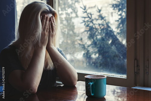 Blonde woman sitting at the table with coffee cup, covering her face with her hands, crying, sad 