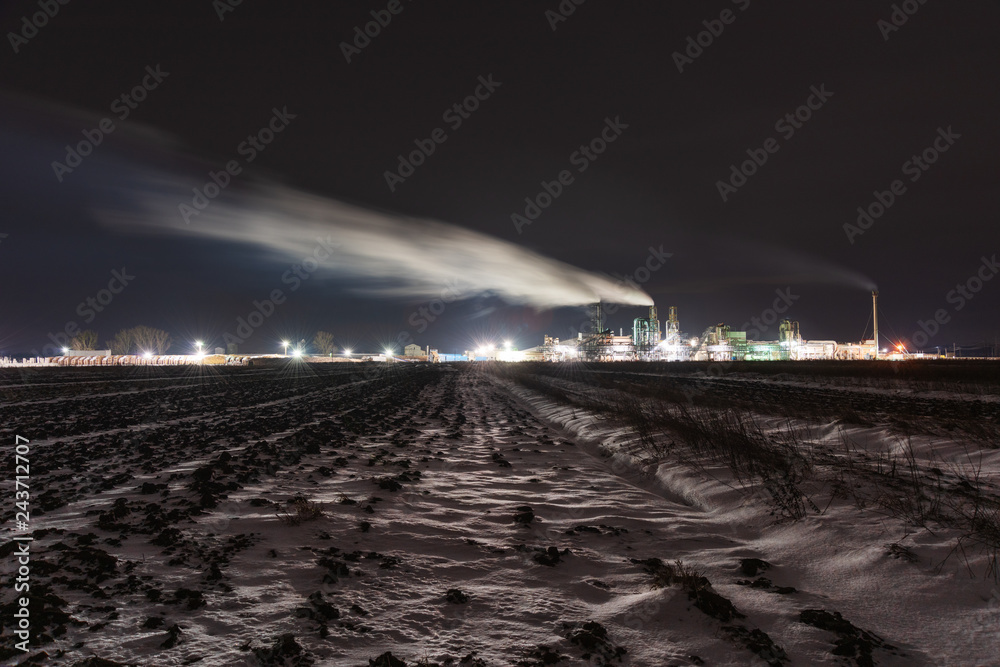 A night view of an industrial unit, a wood-processing plant with fuming pipes and a high-illuminated area on the background of a snow-covered field and a bicycle with a red light.