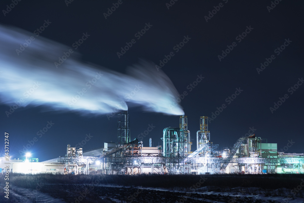 Fototapeta premium A night view of an industrial unit, a wood-processing plant with fuming pipes and a high-illuminated area on the background of a snow-covered field and a bicycle with a red light.