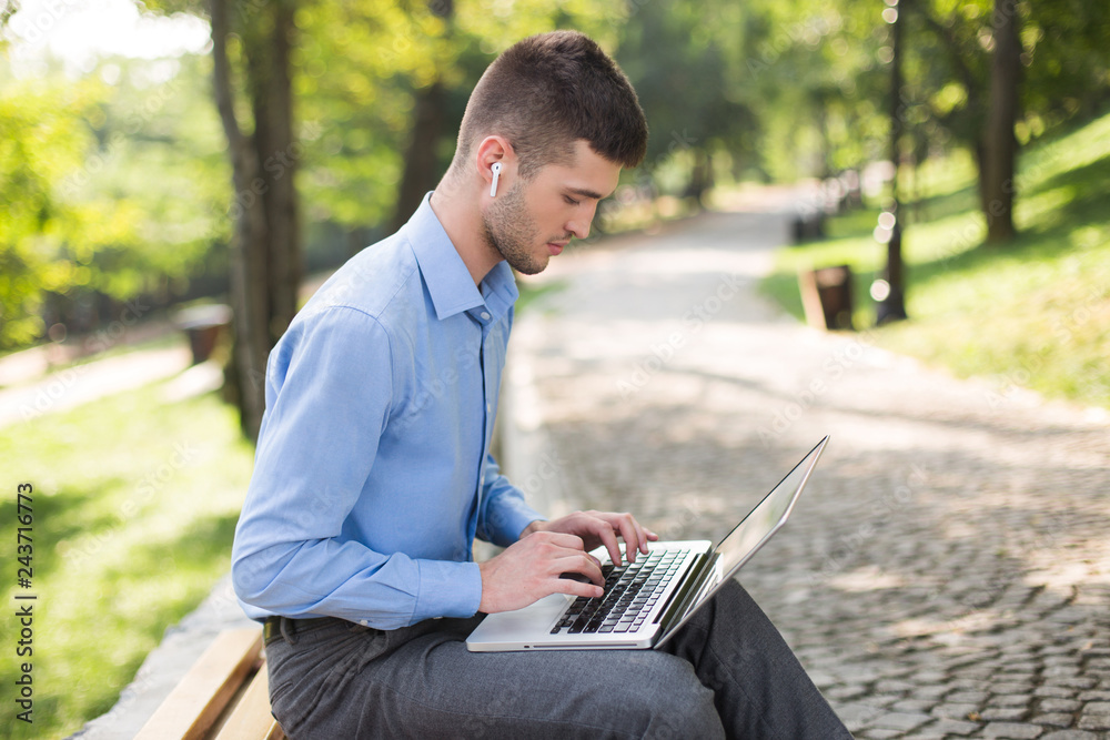 Young handsome man in blue shirt with wireless earphones sitting on bench thoughtfully working on laptop while spending time in park