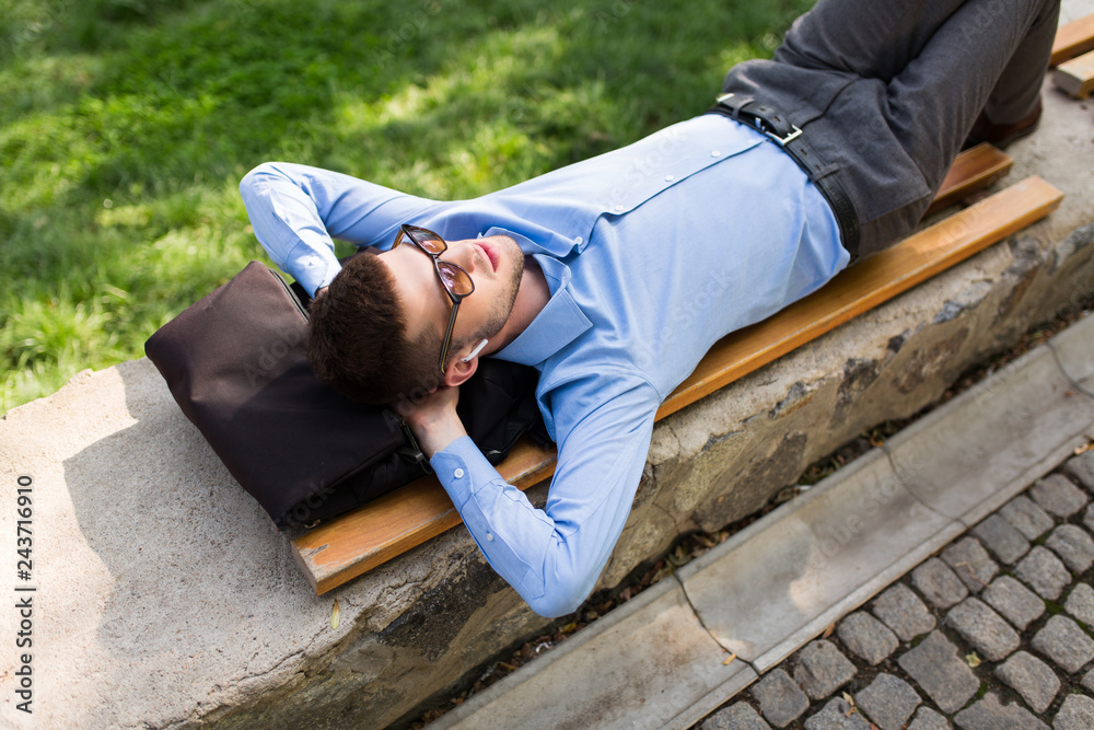 Young handsome man in blue shirt and sunglasses with wireless earphones dreamily lying on bench while spending time in city park Young handsome man in blue shirt and sunglasses with wireless earphones dreamily lying on bench while spending time in city park