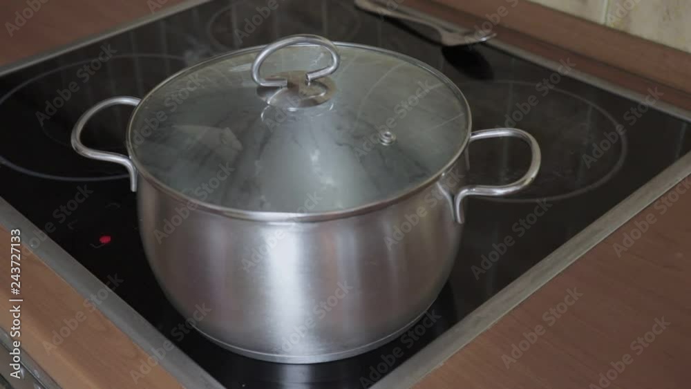 A hand taking off a hot lid from a pan with boiling water on induction glass ceramic stove cook top, the steam goes up A hand taking off a hot lid from a pan with boiling water on induction glass ceramic stove cook top, the steam goes up