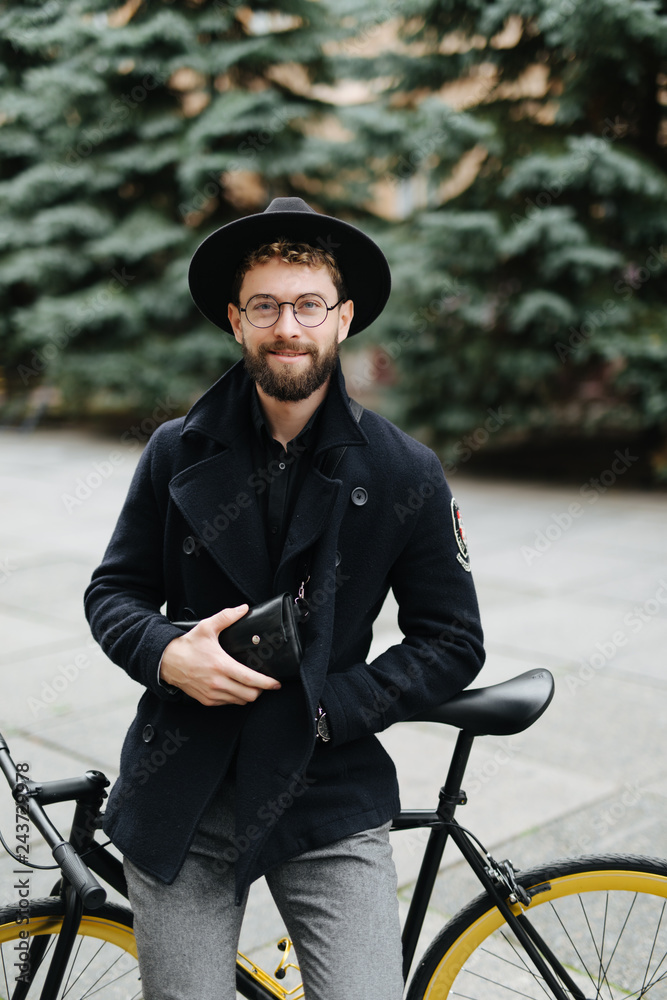Portrait of handsome young bearded man in casual clothes looking away and smiling while leaning on his bike standing outdoors Portrait of handsome young bearded man in casual clothes looking away and smiling while leaning on his bike standing outdoors
