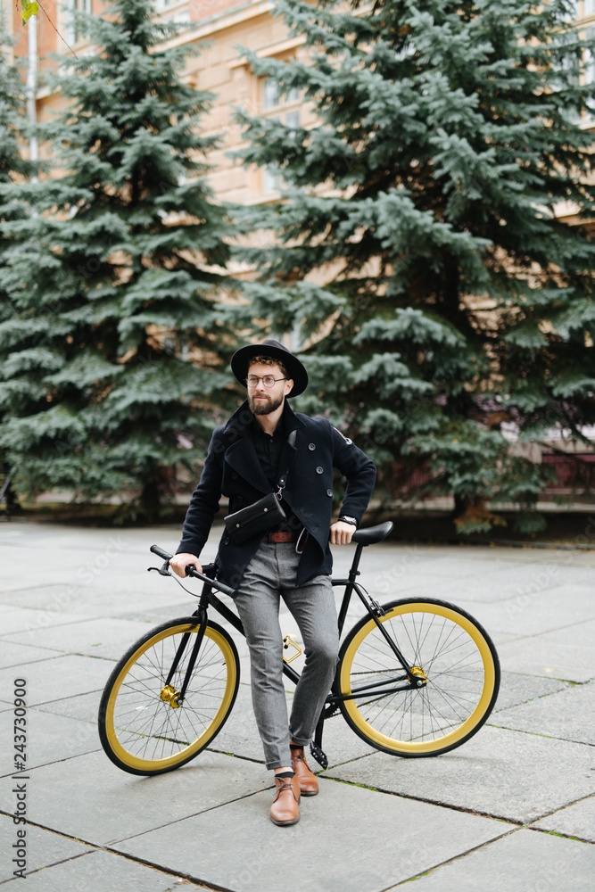 Portrait of handsome young bearded man in casual clothes looking away and smiling while leaning on his bike standing outdoors