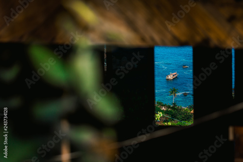  Scenic ocean view framed by a wooden window in a tropical cafe on Koh Tao island, Thailand. Traditional longtail boat floating on blue sea, palm tree and lush greenery in the background.