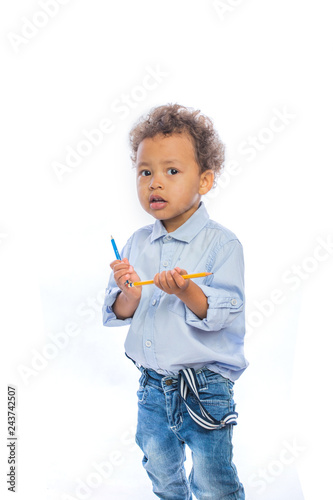 Boy with curly hair in jeans and a light-colored shirt is standing in a half-turn holding a pencils in both hands and looking to the side