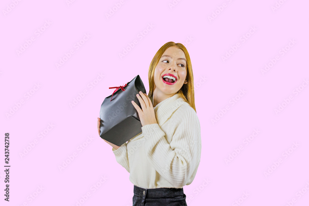 Fototapeta premium A portrait of a beautiful cute red-haired girl who holds a gift bag in a black box with a red velvet ribbon raising a gift, demonstrates his smile, looking enthusiastically to the side against an