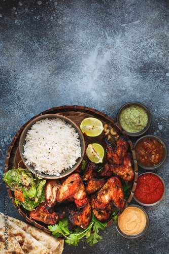 Smoky, roasted tandoori chicken wings served with pilau rice , soft garlic cheese naan with different chutney dipping sauces. Top view, blank space, rustic background