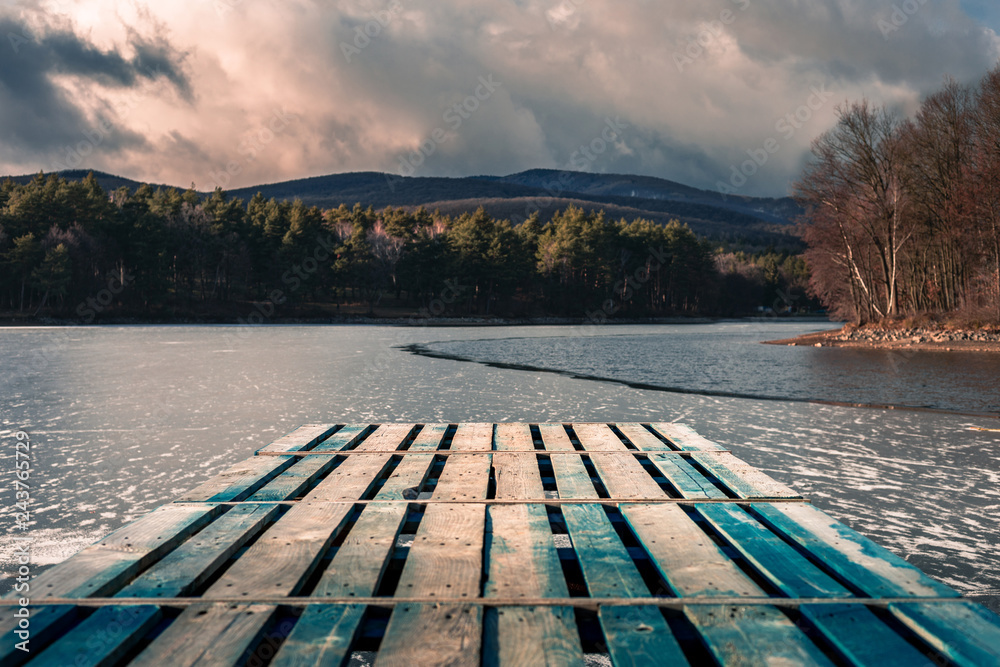 Fototapeta premium Mole (pier) on the lake. Wooden bridge in forest in winter time with blue frozen lake. Lake for fishing with pier. Dark lake (ice) with hills