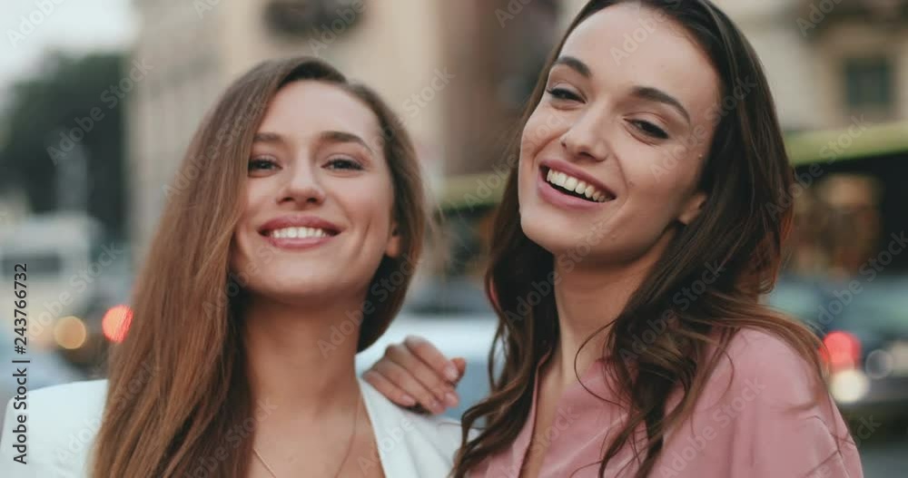 Portrait of the two young Caucasian good looking women, best friends, smiling happily tothe camera while posing at the street with many cars background. Close up. Portrait of the two young Caucasian good looking women, best friends, smiling happily tothe camera while posing at the street with many cars background. Close up.