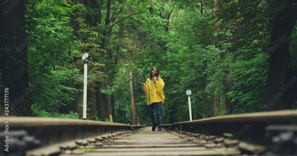 Young Caucasian girl in the raincoat stepping on the railway and getting closer to the camera while walking in the forest. Young Caucasian girl in the raincoat stepping on the railway and getting closer to the camera while walking in the forest.