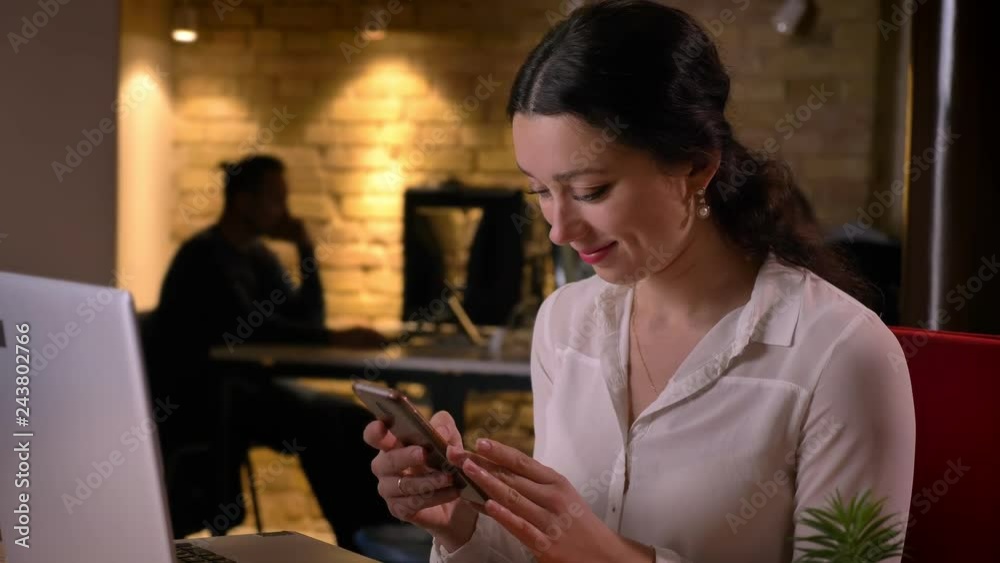 Closeup portrait of young caucasian female office worker browsing on the phone in front of the laptop smiling happily Closeup portrait of young caucasian female office worker browsing on the phone in front of the laptop smiling happily