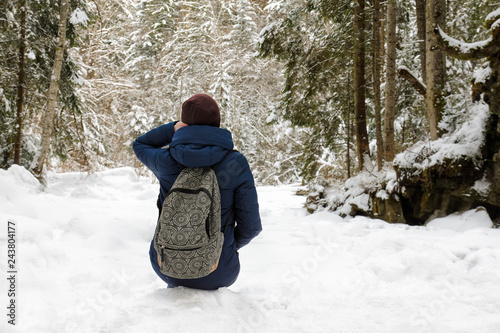 Girl with backpack sitting in a snow-covered coniferous forest. Winter cloudy day.