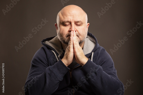 Bald middle-aged man with closed eyes folded hands in prayer, dark background