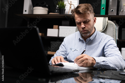 young man sitting at computer table and makes notes in diary