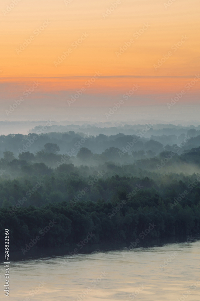 Mystical view on riverbank of large island with forest under haze at early morning. Mist among layers from tree silhouettes under predawn sky. Morning calm atmospheric landscape of majestic nature.