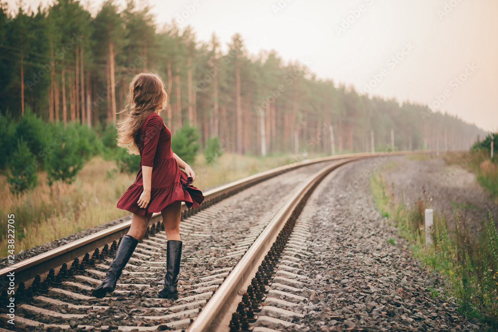 Beautiful dancing girl with curly natural hair enjoy nature in forest on railway. Dreamer lady in burgundy dress walk on railroad. Back view on inspired girl on rails at dawn. Sun in hair in autumn. Beautiful dancing girl with curly natural hair enjoy nature in forest on railway. Dreamer lady in burgundy dress walk on railroad. Back view on inspired girl on rails at dawn. Sun in hair in autumn.