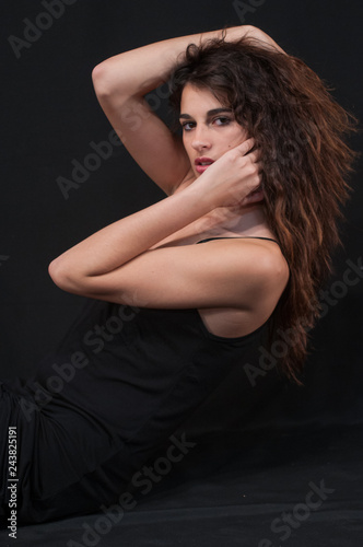 Attractive brunette girl with long wavy brown hair, and perfect makeup, wearing black petticoat dress with thin straps. Fashion model posing on black uniform background.