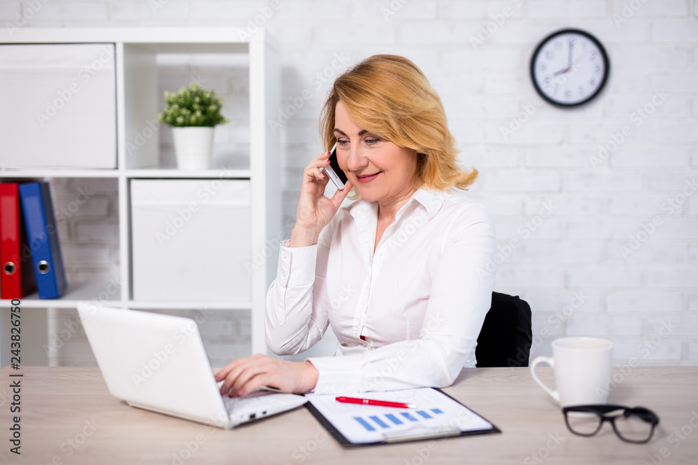 cheerful mature business woman sitting in office and talking by phone