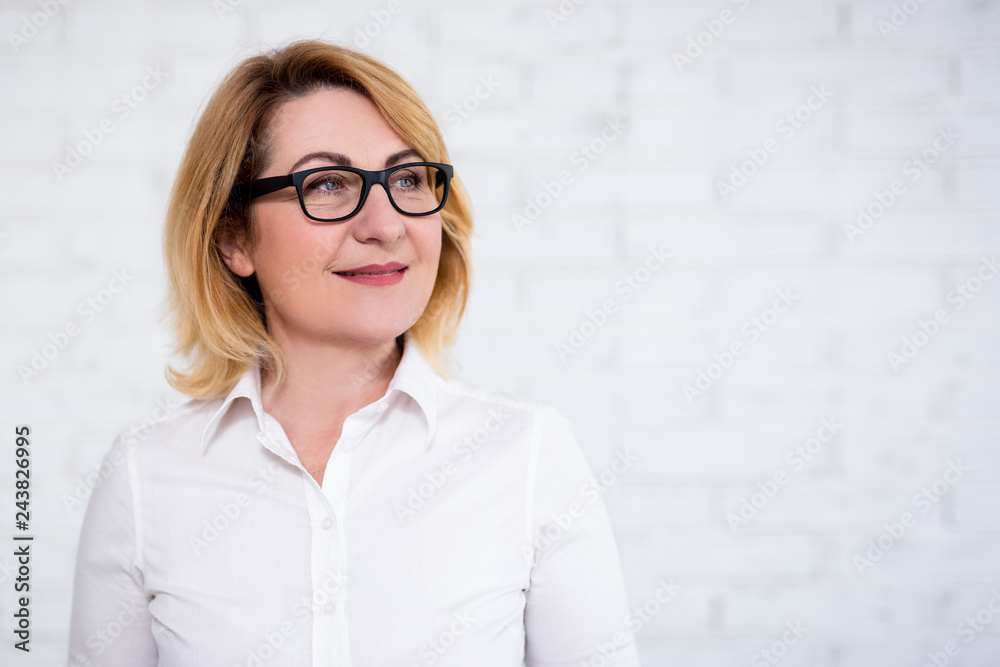 portrait of thoughtful cheerful mature business woman in eyeglasses posing over white wall
