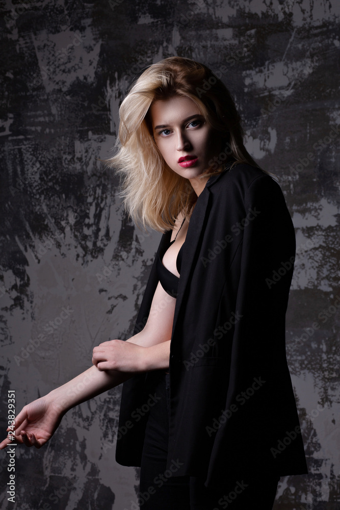 Gorgeous blonde woman with perfect makeup wearing black jacket and bra posing at studio with shadows