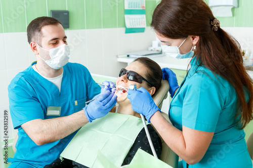 people  medicine  stomatology and health care concept - male dentist and assistant with dental curing light and mirror treating woman patient teeth at the real dental clinic office