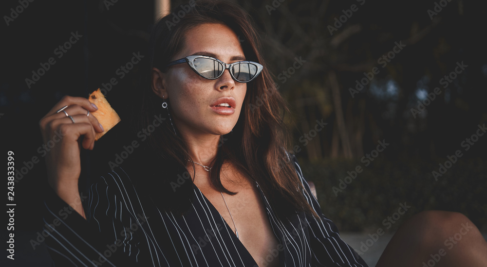 Outdoor fashion photo of young beautiful lady in sunglasses at villa. sexy woman holding piece of pineapple. Summer vibes