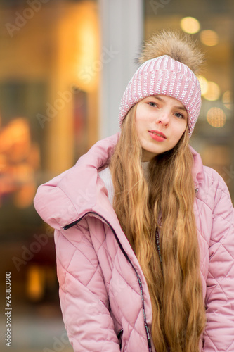 A girl in a pink warm jacket and hat with .pompon stands on the background of golden yellow lights and blurred shop windows. Winter wonderland. Christmas and New Year concept.