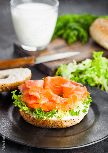 Fresh healthy bagel sandwich with salmon, ricotta and lettuce on black plate on black kitchen table background. Healthy diet food. Glass of milk and fresh vegetables
